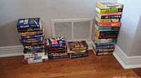 Stack of paperback novels stacked against a wall, titles visible including Tom Clancy, Robert Ludlum, John Grisham, and other authors
