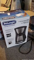 Boxed DeLonghi drip coffee maker showing brand and model details on the packaging.