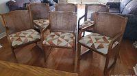 Overall view of seven wooden dining chairs with cane backs and geometric patterned padded seats arranged together showing armrests on some chairs and cane backs.