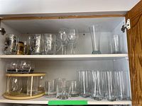 Two white shelves filled with various clear drinking glasses including beer mugs, wine glasses, and tumblers inside a cabinet.