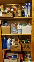 Four shelves of wooden shelving filled with assorted household cleaning products, food prep items, and plastic containers organized in boxes and loose arrangement.