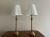 Front view of the pair of brass-tone metal framed lamps with white pleated shades on a wood surface against a beige wall.