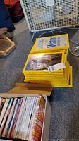 Stack of National Geographic magazines with their distinctive yellow border covers, placed on carpeted floor next to a box containing other magazines.