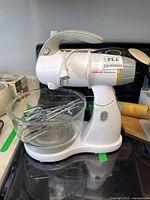 Sunbeam Mixmaster stand mixer on a stove with clear glass mixing bowl and two metal beaters inside the bowl, power cord and plug visible.