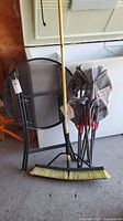 Front view of black metal mesh round folding patio table, folding camp chair and yellow push broom leaning on them.