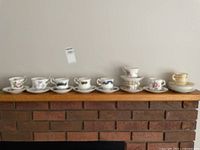 Eight teacups placed on saucers arranged in a row on a mantle, showing varied floral patterns and colors, some with gold rim accents.