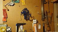 Wooden pegboard with various household tools including a red staple gun, metal hand planer, metal clamps, paint brushes, measuring tape, wire cutters, and assorted hooks. Some tools are in original packaging.