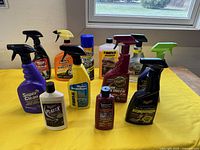 Eleven assorted automotive cleaning and detailing products displayed on wooden table under window. Spray bottles and plastic bottles with labels showing various brands including Meguiar's, Rain-X, Tanner's Preserve.