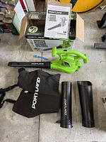 Full view of the Portland 3-in-1 blower/vacuum/mulcher with two black attachment tubes, large black collection bag with 'PORTLAND' text, instruction manual, and original box on a concrete floor.