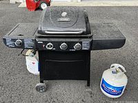 Front view of black Thermos propane grill with hinged lid and three control knobs. One control knob is broken. Two propane tanks visible: one connected to grill and one nearby.