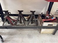 Five automotive jack stands displayed in a row on a metal shelf, including one red painted stand and four unpainted metal stands with rust.