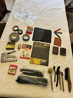 Photo showing assortment of sandpapers, zip ties, stapler, wire brushes, and other small hand tools on a white surface.