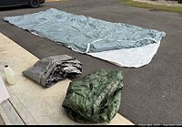 Lot laid out on driveway showing three tarps: large gray and white zippered car pocket cover spread out, folded silver tarp and green camouflage tarp with grommets folded on the side.