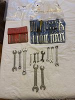 Wrenches arranged loosely on white sheet, including various sizes and types. Some in blue plastic holder and red pouch.