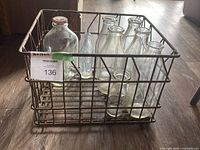 Photo showing front side of metal antique milk crate holding assorted glass milk bottles on wooden floor.