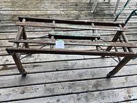 Full side view showing rectangular wooden frame with four legs, cross supports, and metal brackets on floor decking outdoors.