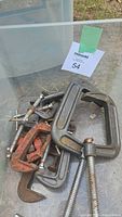 Bin of assorted C-clamps showing multiple clamps of different sizes, some red painted, some plain metal with rust.