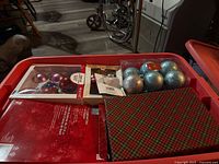 Wide view of plastic tote filled with Christmas decorations including boxes of glass ball ornaments in red, purple, and silver, Christmas cards with plaid design, and packaging for star tree topper.