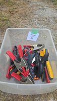 Top view of assorted clamps with red, yellow, and black handles in a plastic bin outdoors on gravel ground.