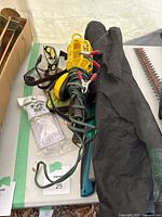 Photo showing miscellaneous gardening tools on a table including yellow work light with power cord, partially unrolled black landscape fabric, and three sets of safety glasses (black, clear, yellow frames) next to some gardening tools.