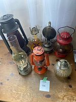 Wide view of six assorted lanterns on a wooden table, showing variety in colors, styles, and materials.
