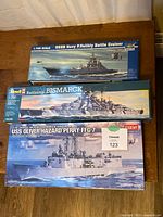 Three sealed model ship kits boxed: USS Navy P. Velikiy Battle Cruiser (Trumpeter), Battleship Bismarck (Revell), USS Oliver Hazard Perry FFG-7 (Academy) arranged on a wooden surface.