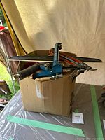 Box filled with caulking tools including a blue caulk gun, red and black handled tools, and other painting accessories.