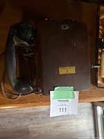 Front view of antique telephone mounted on wooden surface showing wooden box, black handset, and attached cord with a label on the wooden case.