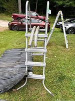 Gray metal pool ladder with curved handrails and five steps standing on grass, with hoses attached.