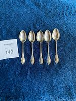 Set of 5 silverplated spoons with decorative handles laid out on a blue textured background. The spoons show tarnish and need cleaning.