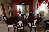 Dining room set with one large dining table and six chairs around it in a dining room with red paint on wall, natural light from window, glass cabinet behind.