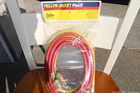 Photo of three coiled refrigeration charging hoses in sealed original packaging, displayed on wooden table with white chair background.