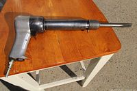 Silver metal heavy duty air chisel with long cylindrical barrel and a flat, wide chisel tip lying on a wooden table, shown from the left side.