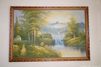 Framed oil painting showing a nature scene with trees, waterfall, river, small house, and mountains in the background.