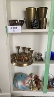 Overview of shelf with various brass items including brass vases, candlesticks, scoops, and decorative figurines along with a painted animal figurine