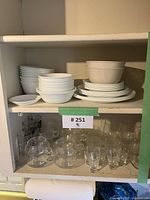 Kitchen cupboard shelf with stacked white Corelle dishes including bowls and plates, and assorted clear glass mugs and glasses below.