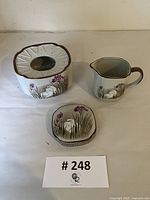 Three porcelain stoneware pieces with speckled finish and hand-painted white cranes and purple iris flowers: rectangular box with lid, small pitcher, and small flat dish.