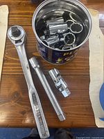 Photo showing 1/2 inch drive ratchet, long extension bar, swivel extension bar and metal can with various sockets inside on wooden surface.