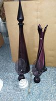 Two tall purple glass items with rounded bases and pointed tops placed on carpet with a cardboard background.