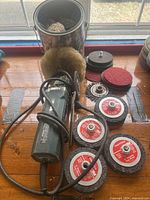 Full view of Metabo hand grinder with several grinding and polishing wheels and wire brush attachment on wooden surface next to a can of buffing wheels.