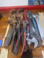 Top view of all tools piled together on a wooden floor, showing rusty pipe wrenches, adjustable wrenches, pliers and utility cutter.