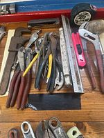 Overview of assorted hand tools on wooden surface including pliers, wrenches, measuring tools, and hammer