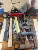 Top-down image showing full set of miscellaneous hand tools on wooden floor including hammers, pliers, wrenches, measuring tapes, pipe cutter, and scraper.