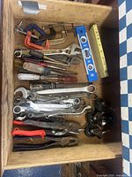 Drawer containing various hand tools including C clamps, adjustable wrenches, screwdrivers, pliers, a level, measuring tape, and casters.