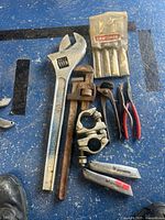 Overview of all tools in the lot displayed on a blue surface showing adjustable wrench, pipe wrench, utility cutters, needle nose pliers, chisel set, and cable clamps.