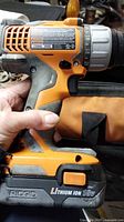 Side view of orange and gray Ridgid 18V lithium ion cordless drill held in hand, showing battery and part of carrying bag.