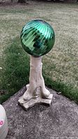 Green glazed glass yard ball mounted on white pedestal resembling animal paws, outdoors on grass.