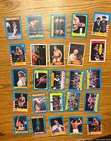 Front sides of 25 vintage wrestling cards laid flat on a wooden surface showing colorful images of wrestlers and match scenes.