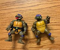 Front view of two Teenage Mutant Ninja Turtle action figures with purple masks, tan plastrons, and articulated limbs on a wooden surface.