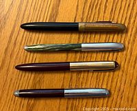 Four vintage ballpoint pens arranged horizontally on a wood grain surface, showing full lengths and varying styles and colors.